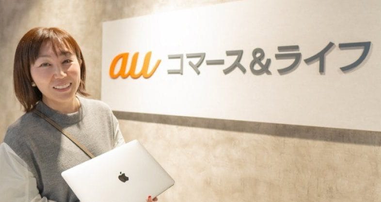 auコマース＆ライフのロゴの前でノートパソコンを持つ担当者